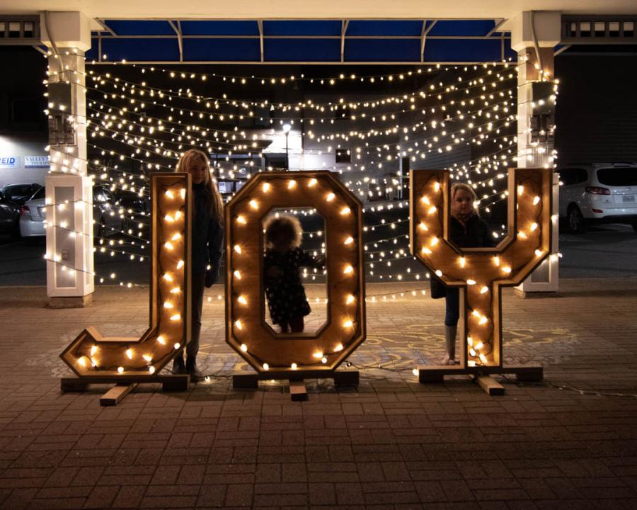 A family of three kids pose behind life-sized wooden letters that spell out the word joy. A backdrop of beautiful lights shines behind them. 