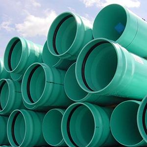 Green sewer pipes are stacked in a pile against a backdrop of blue sky and fluffy white clouds