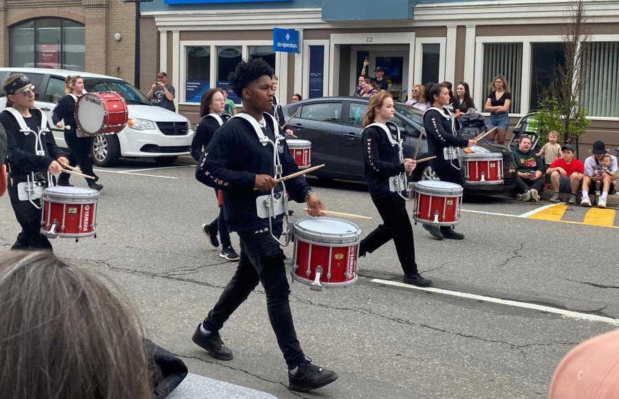 A youth in band uniform marches with a group of musicians down webster street in kentville carrying a snare drum on his hip and holding drumsticks