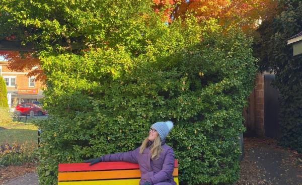 A woman sits on a rainbow coloured bench in front of a gorgeous japanese maple tree that is turning bright orange with the fall temperatures