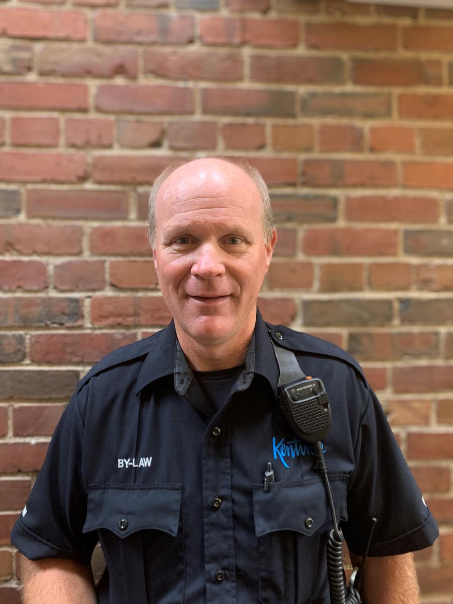 A white man with thinning hair is wearing a bylaw enforcement uniform against a brick wall backdrop
